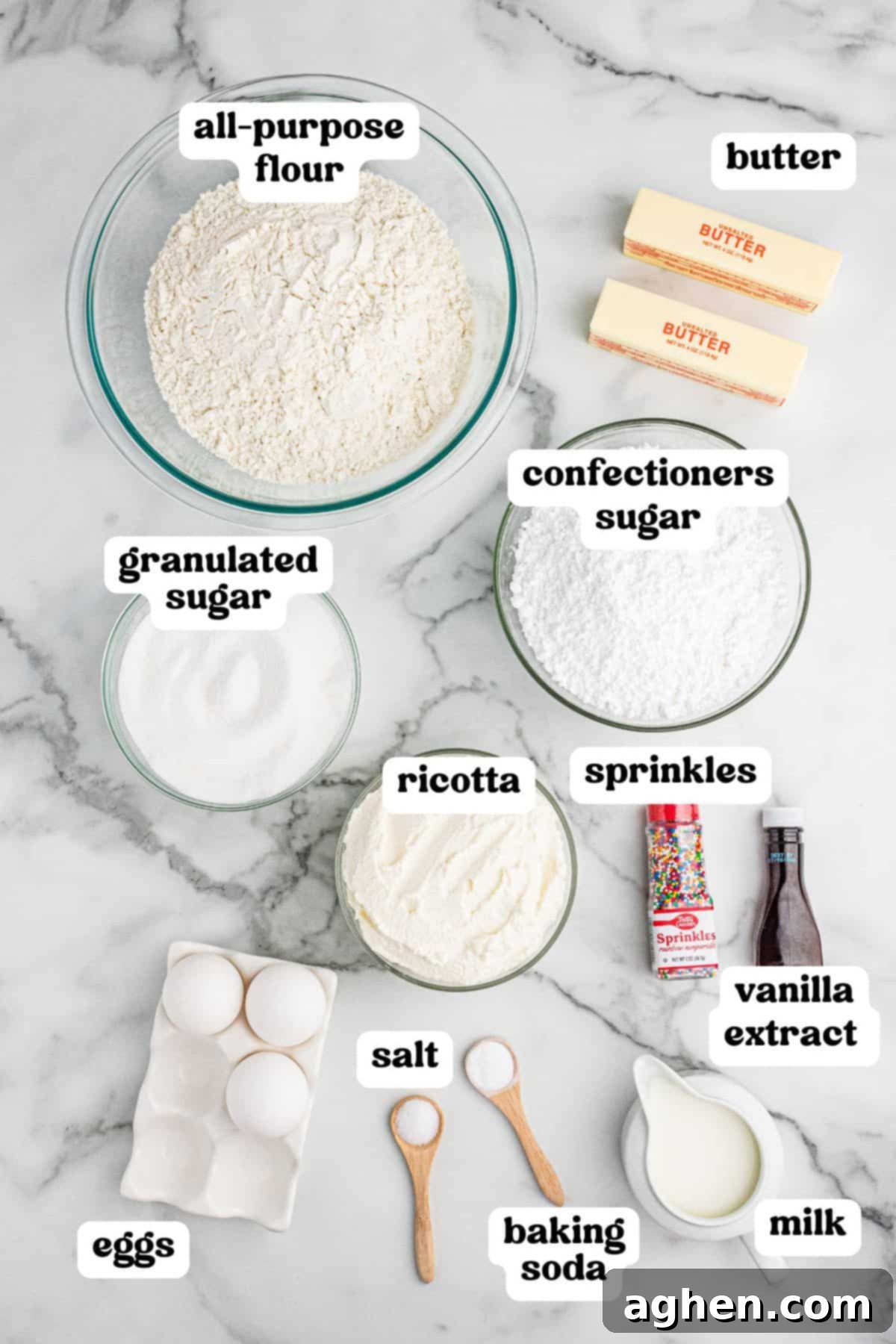A top-down view of ricotta cookie ingredients labeled with text. The image includes bowls of all-purpose flour, granulated sugar, confectioners sugar, and ricotta, along with sticks of butter, eggs, salt, baking soda, milk, vanilla extract, and colorful sprinkles, all neatly arranged on a marble countertop.