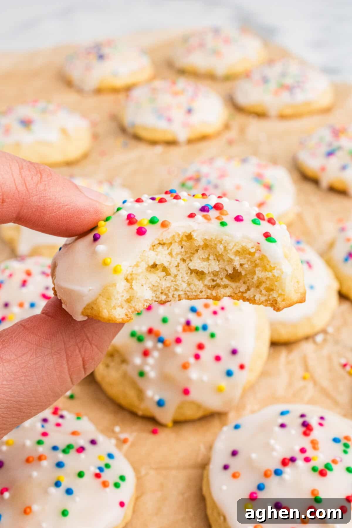 A hand holds a half-eaten Italian ricotta cookie with white icing and colorful sprinkles, revealing its soft, fluffy interior. More cookies are scattered in the background on parchment paper, highlighting the festive and homemade presentation.