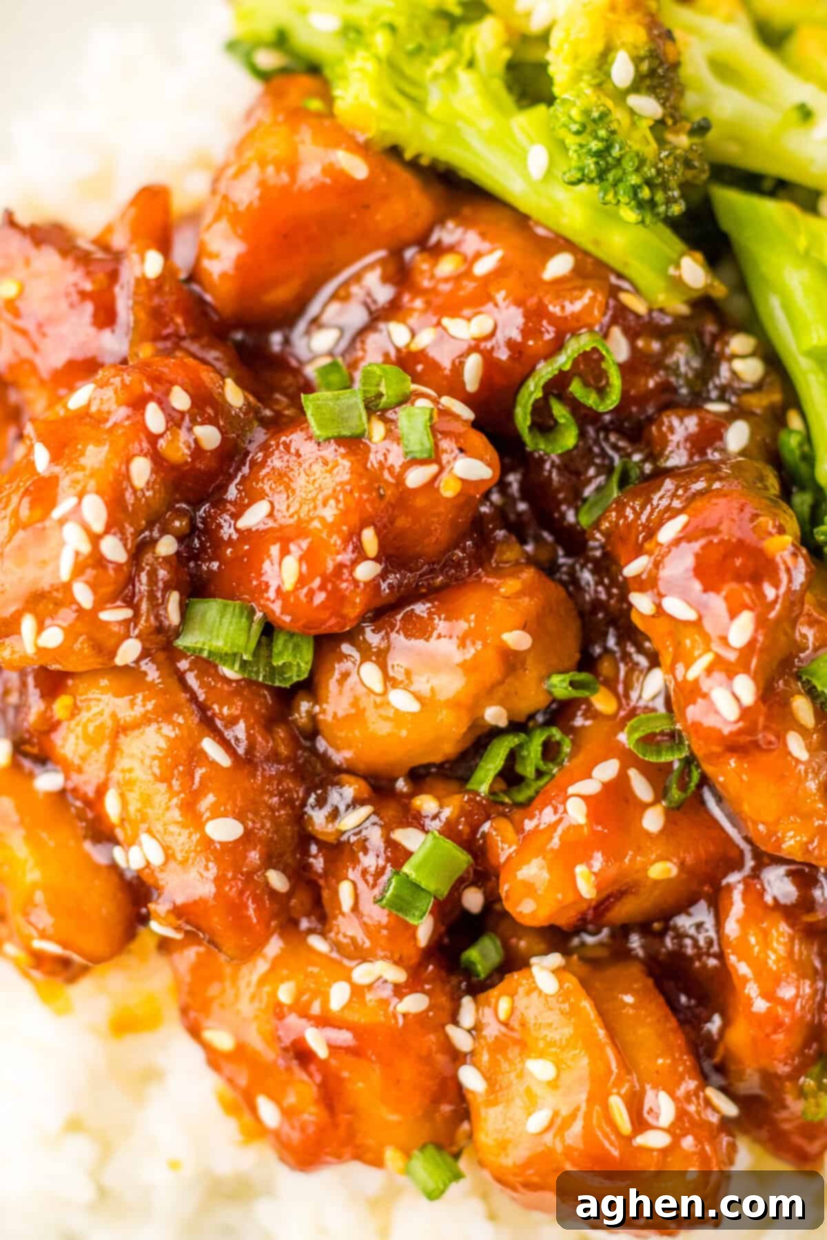 A close-up of copycat Chinese bourbon chicken coated in a rich glaze and sprinkled with sesame seeds and chopped green onions. The chicken pieces are tender and caramelized, sitting on a bed of rice with steamed broccoli visible in the background. 