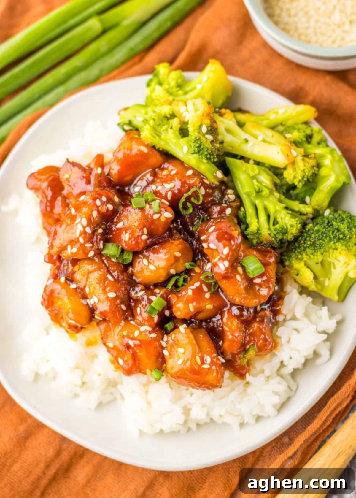 A plate of mall food court style bourbon chicken with sesame seeds and green onions, served with steamed broccoli on a bed of white rice.