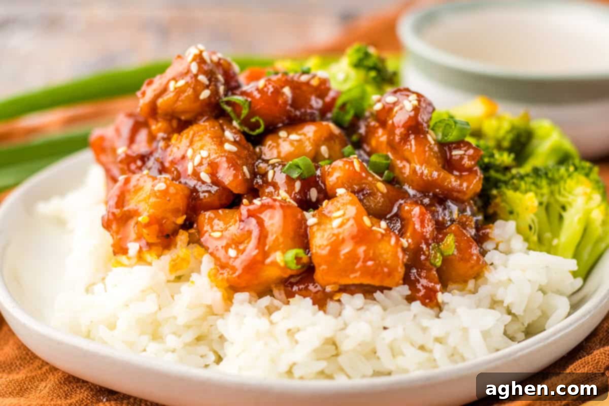 A generous serving of bourbon chicken, coated in a shiny, dark glaze and sprinkled with white sesame seeds and chopped green onions. The mall food court style chicken dish is served over a bed or white rice and with a side of broccoli.