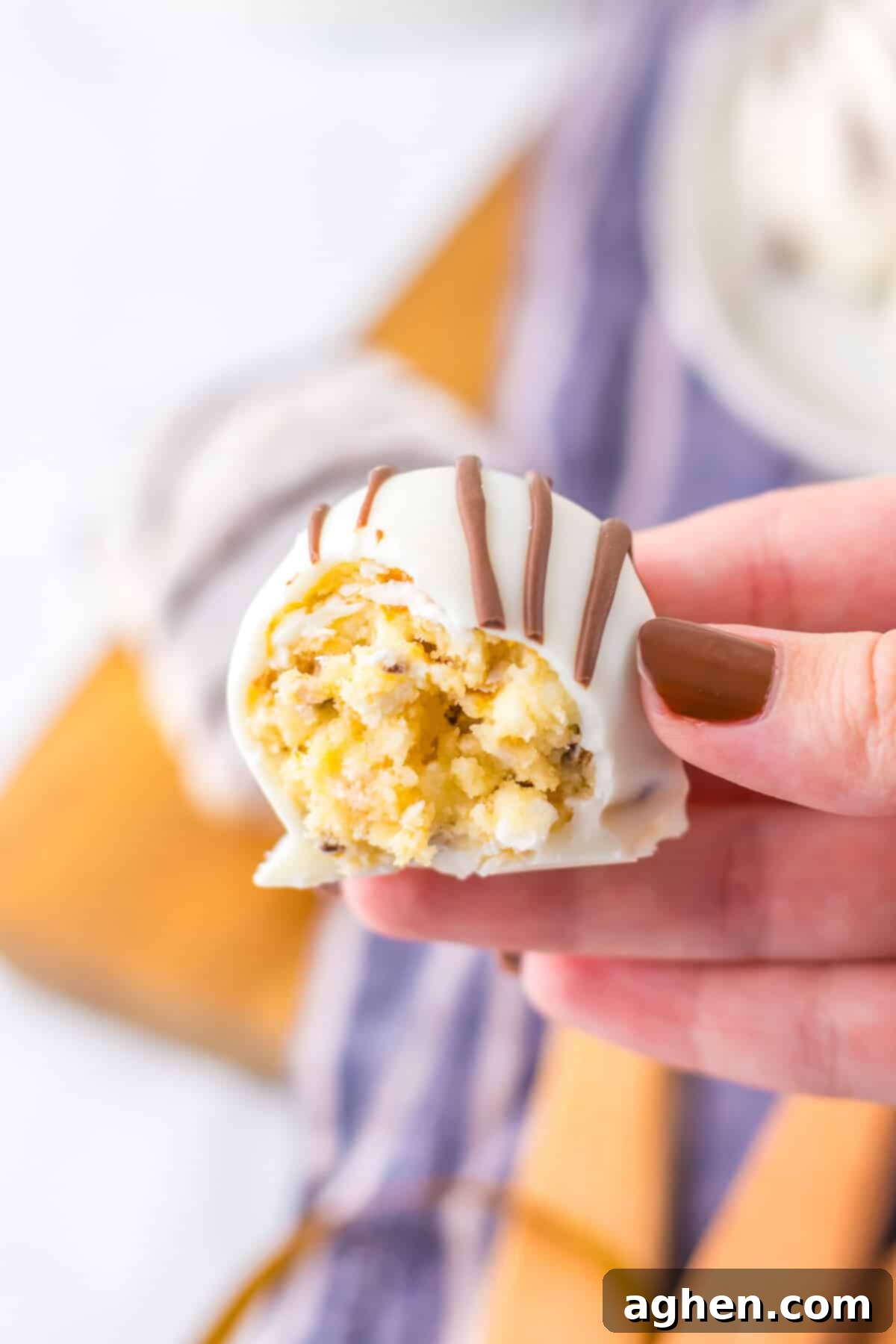 A hand holding a white chocolate-coated truffle (Zebra Cake Truffle) with a bite taken, showing the yelllow cream cheese and cake filling inside.