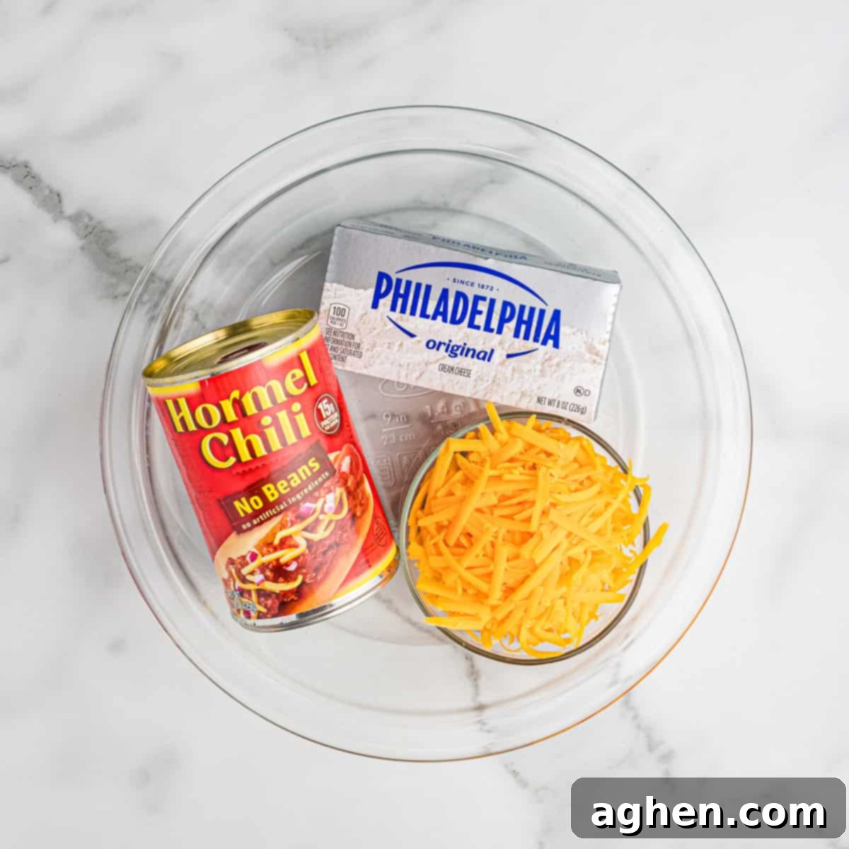 Ingredients for 3-ingredient chili cheese dip displayed in a glass pie dish: a block of Philadelphia cream cheese, a can of Hormel chili (no beans), and shredded cheddar cheese. 