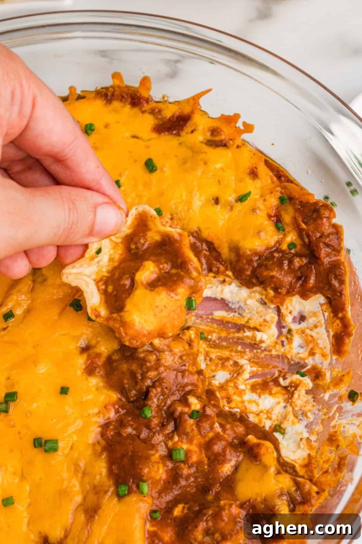 A hand dipping a tortilla chip into the partially eaten chili cheese dip, showing the creamy layers and melted cheese.