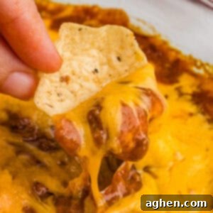 A close-up of a tortilla chip being dipped into the hot, melted chili cheese dip, showing the rich layers of melted cheddar and chili.