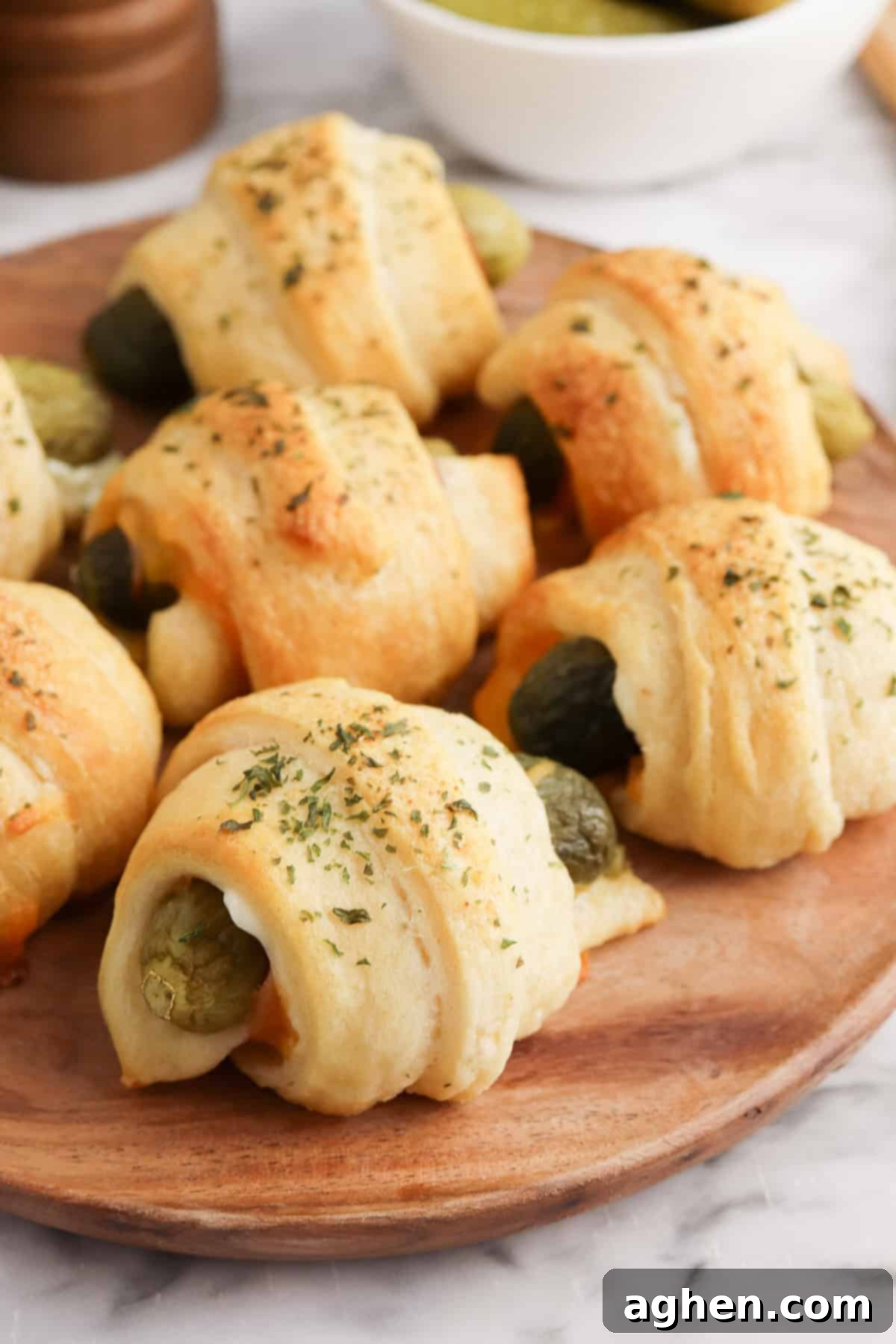Golden-brown crescent roll-wrapped pickles sprinkled with dried herbs are arranged on a round wooden plate. Melted cheese is visible inside some of the rolls.