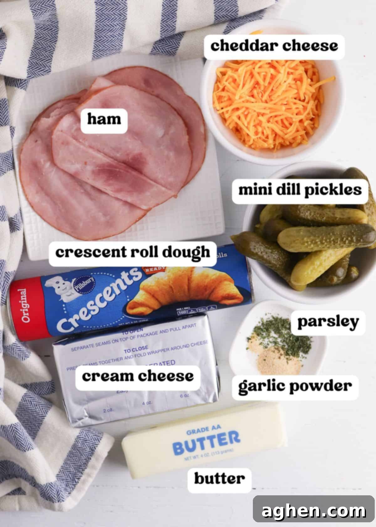 A flat lay of ingredients for pickles in a blanket with text labels. Visible items include slices of ham on a white plate, shredded cheddar cheese in a white bowl, a can of crescent roll dough, a block of cream cheese, mini dill pickles in a bowl, butter, and a small dish with parsley and garlic powder