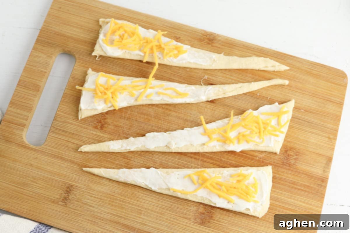 Four triangular pieces of crescent roll dough are laid out on a wooden cutting board. Each triangle is evenly spread with cream cheese and topped with scattered shredded cheddar cheese.