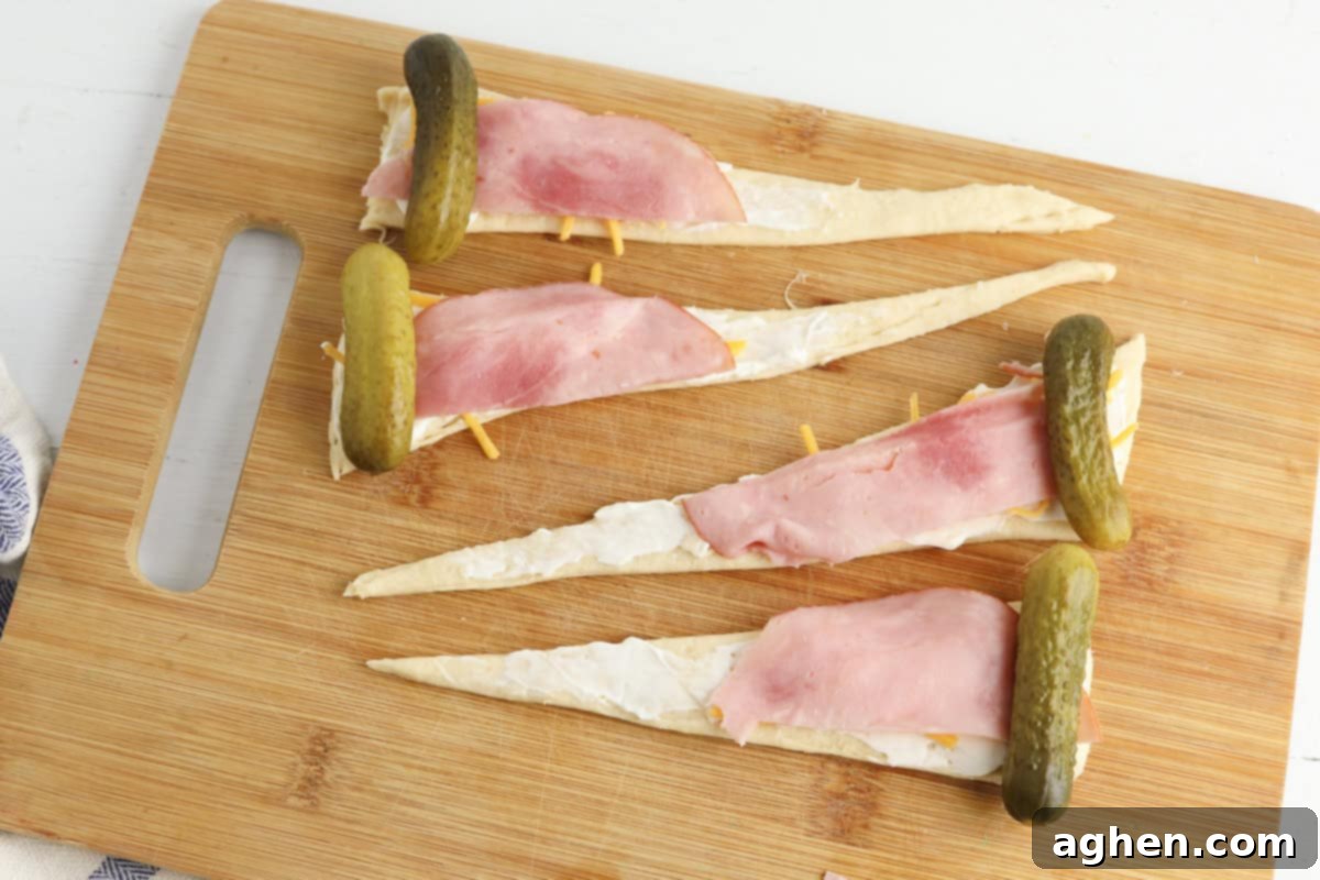 Each triangle is spread with cream cheese, topped with shredded cheddar, a slice of ham, and a whole pickle placed at the widest end.