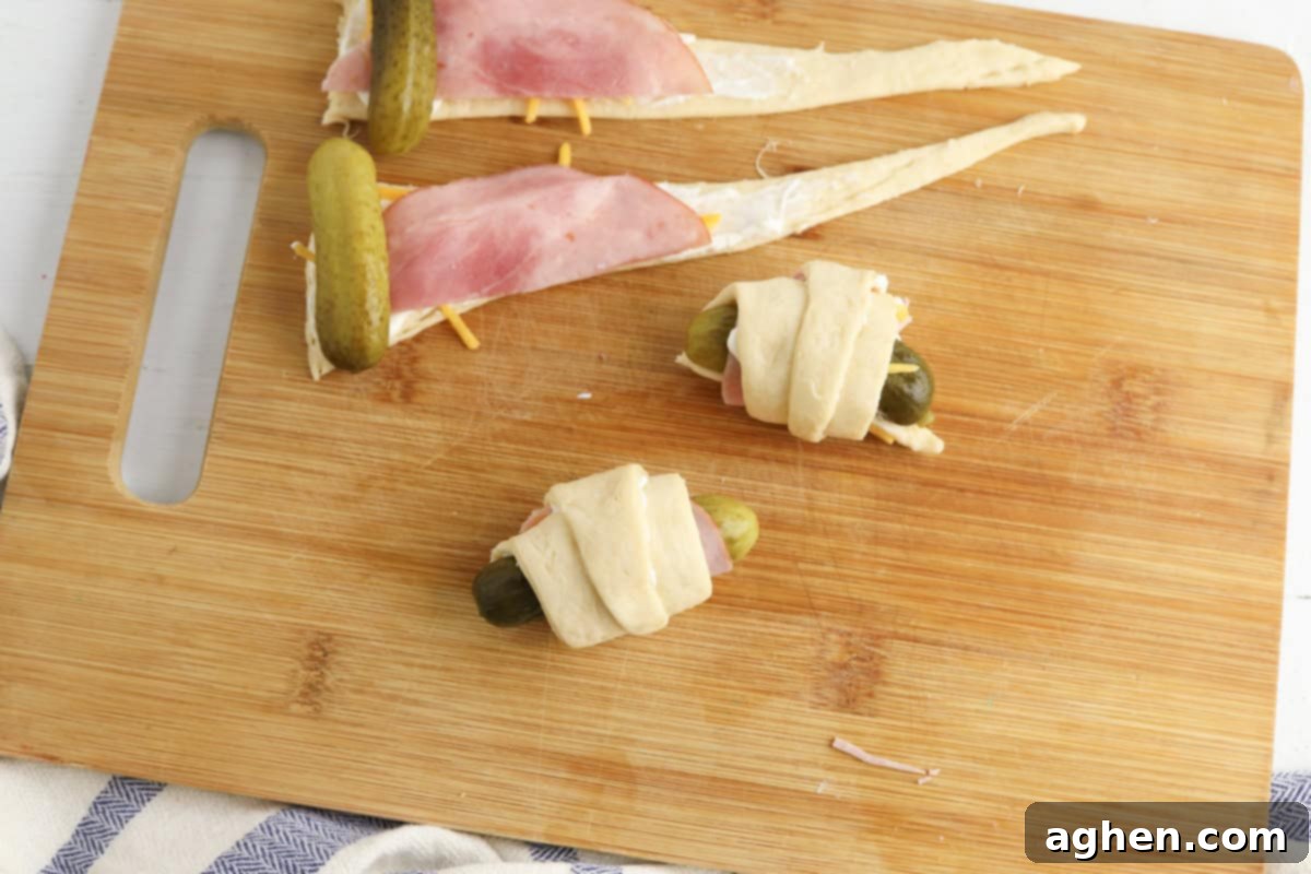 Two completed pickles wrapped in crescent roll dough next to two unwrapped triangles of dough layered with cream cheese, shredded cheddar, ham, and pickles.