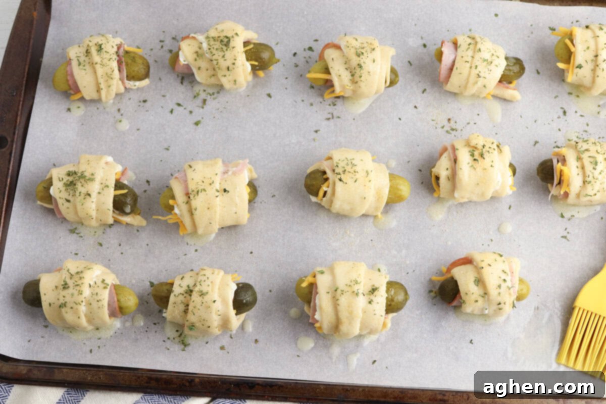 Pickles wrapped in crescent roll dough are evenly spaced on a parchment-lined baking sheet. The dough is topped with melted butter and sprinkled with herbs.