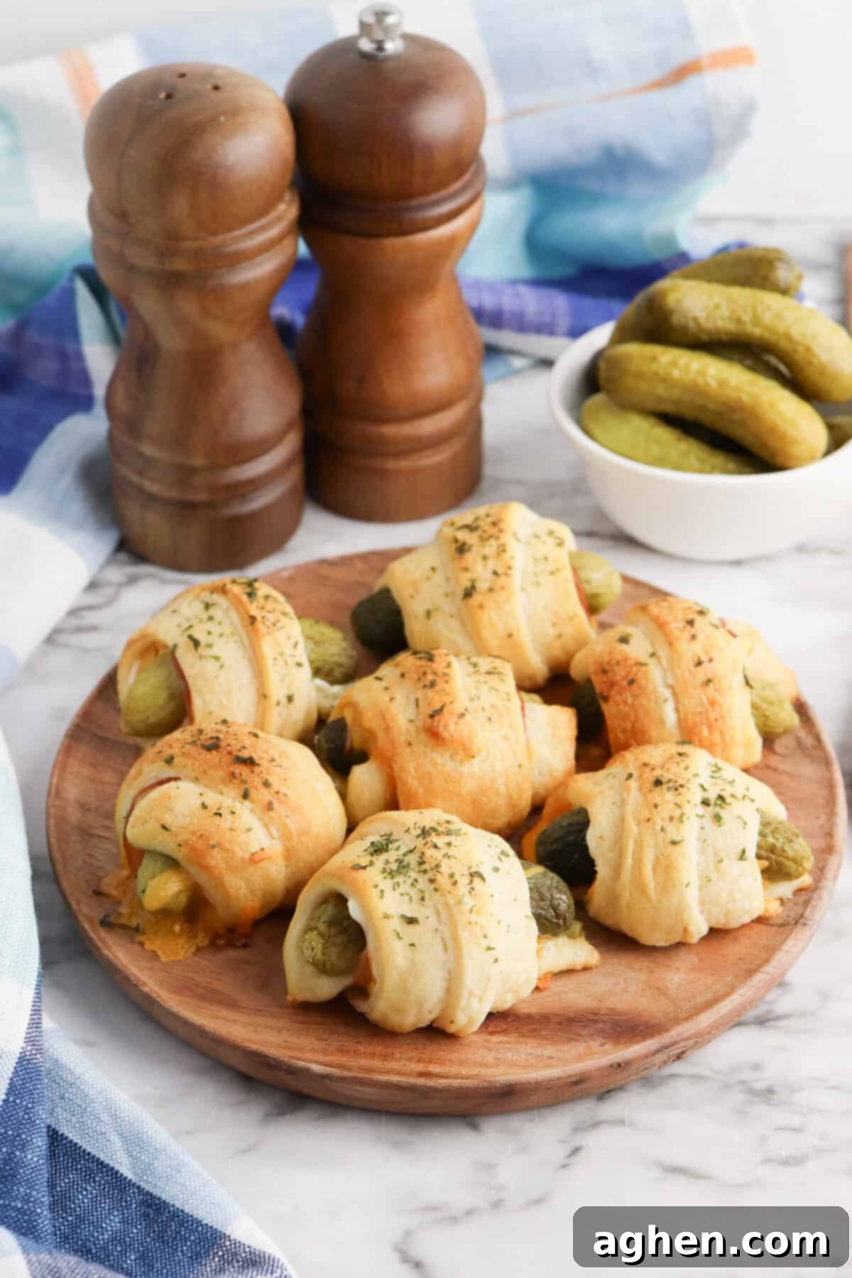 Pickles in a blanket on a wooden plate with melted cheese visible and bowl of mini dill pickles in the background.