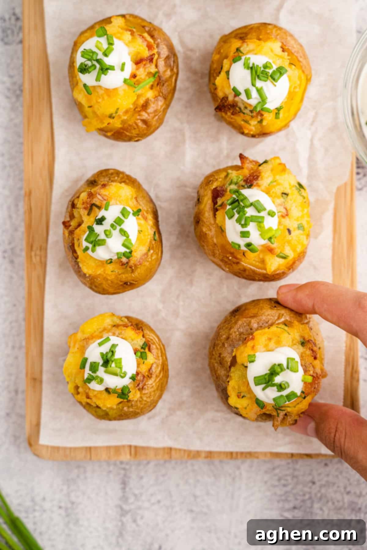 Overhead view of six perfectly prepared twice-baked mini potatoes arranged on parchment paper, beautifully garnished with cool sour cream and fresh chopped chives, with a hand reaching in to grab one of the delicious appetizers.