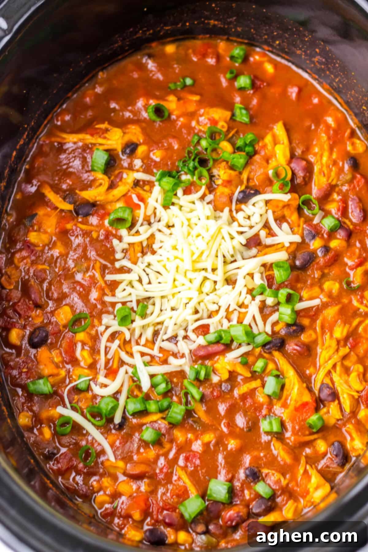 An overhead view of slow cooker taco soup with chicken inside a black crockpot. The soup is topped with shredded cheese and chopped green onions, featuring visible ingredients like shredded chicken, black beans, corn, and diced tomatoes in a hearty broth.