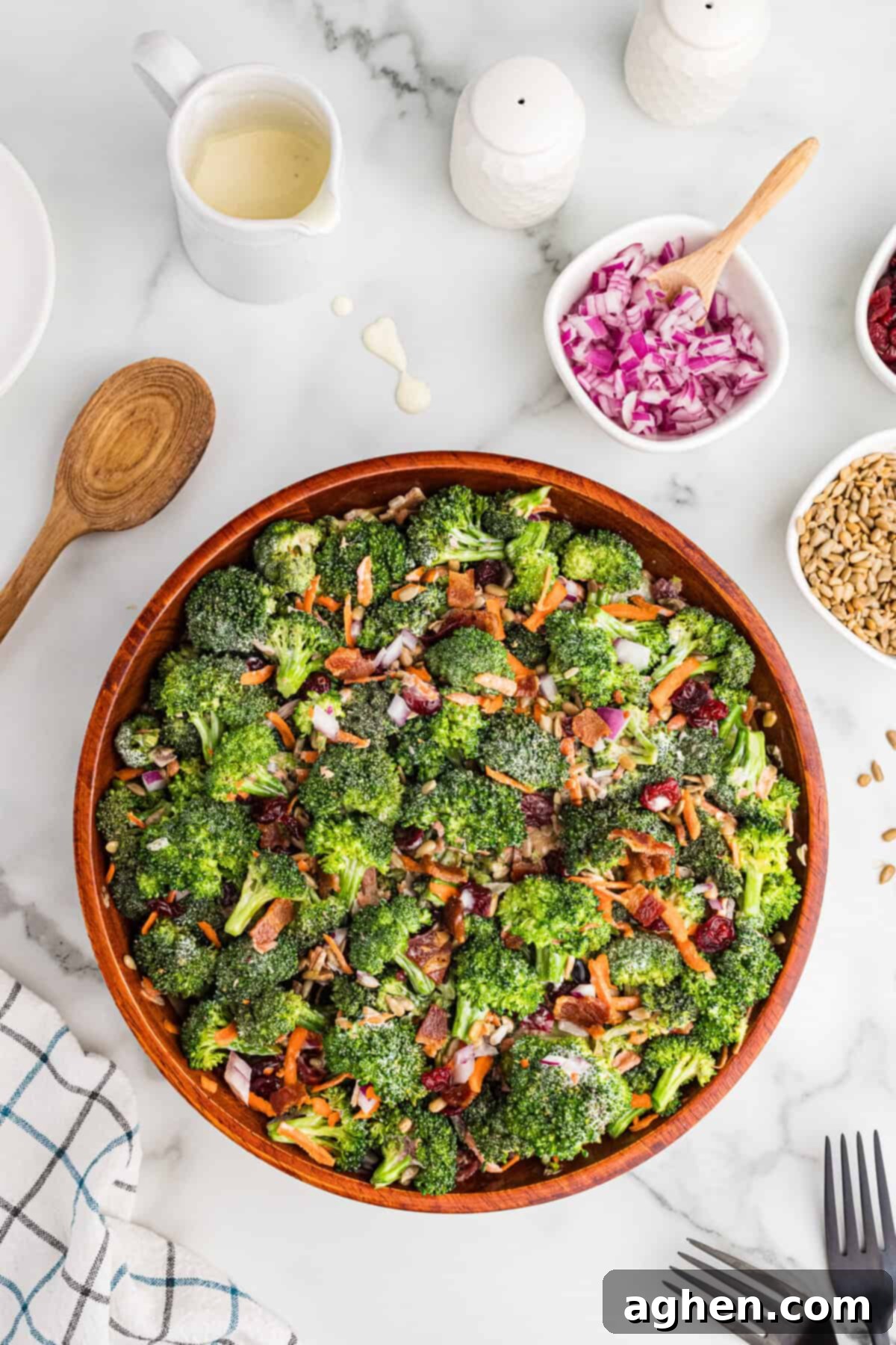 Super Crunchy Broccoli Delight 7 A top-down view of a large wooden bowl filled with broccoli crunch salad, featuring broccoli, bacon, cranberries, shredded carrots, sunflower seeds, and red onions. The bowl is surrounded by small dishes of extra ingredients, a wooden spoon, and a checkered kitchen towel on a marble surface.