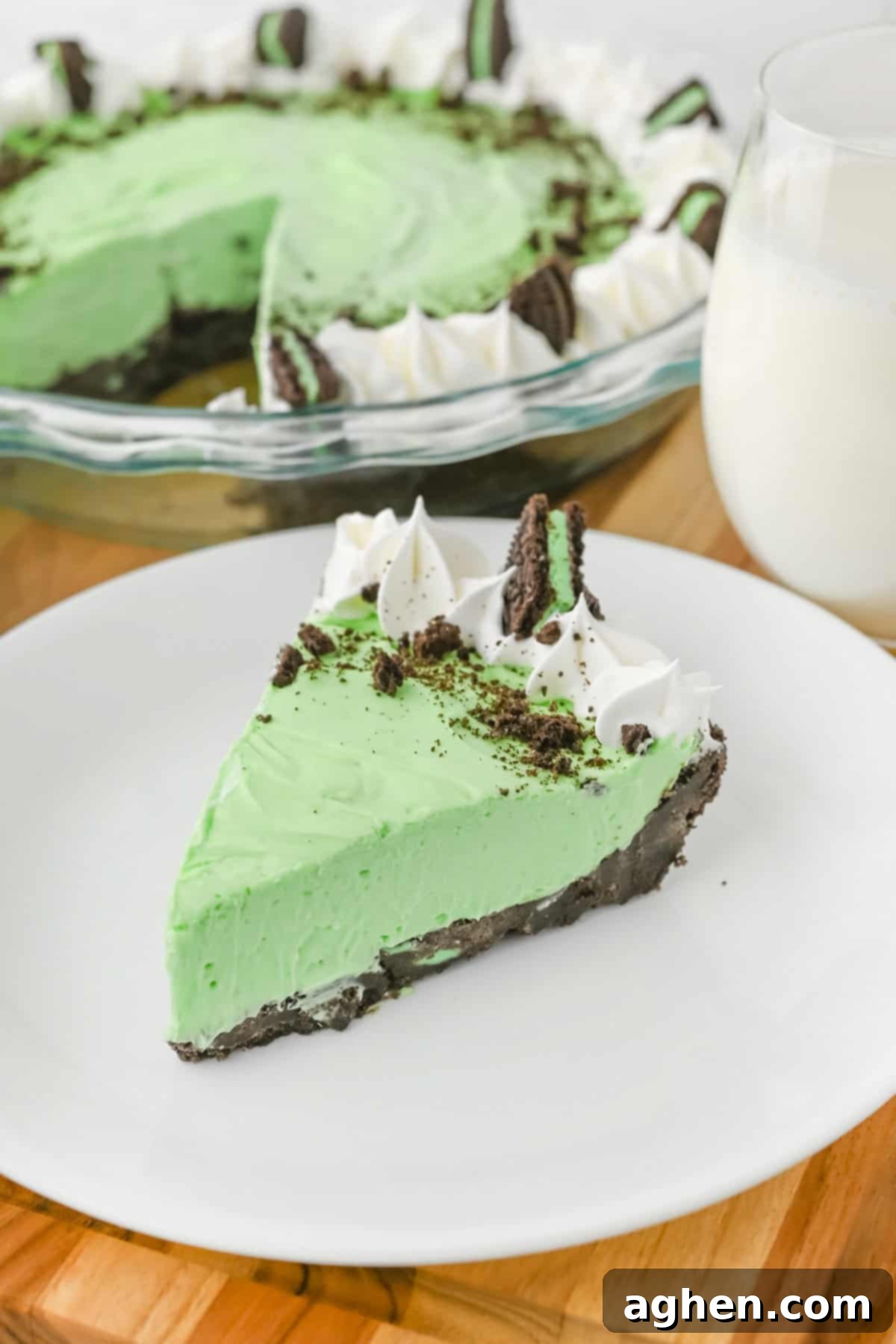 A perfectly sliced piece of no bake grasshopper pie on a white plate, showcasing its smooth, vibrant green mint filling and dark chocolate cookie crust. It's elegantly topped with a swirl of whipped cream, a sprinkle of crushed chocolate cookies, and a whole Mint Oreo piece. In the soft-focus background, the full pie in a glass dish and a glass of milk complete the inviting scene.