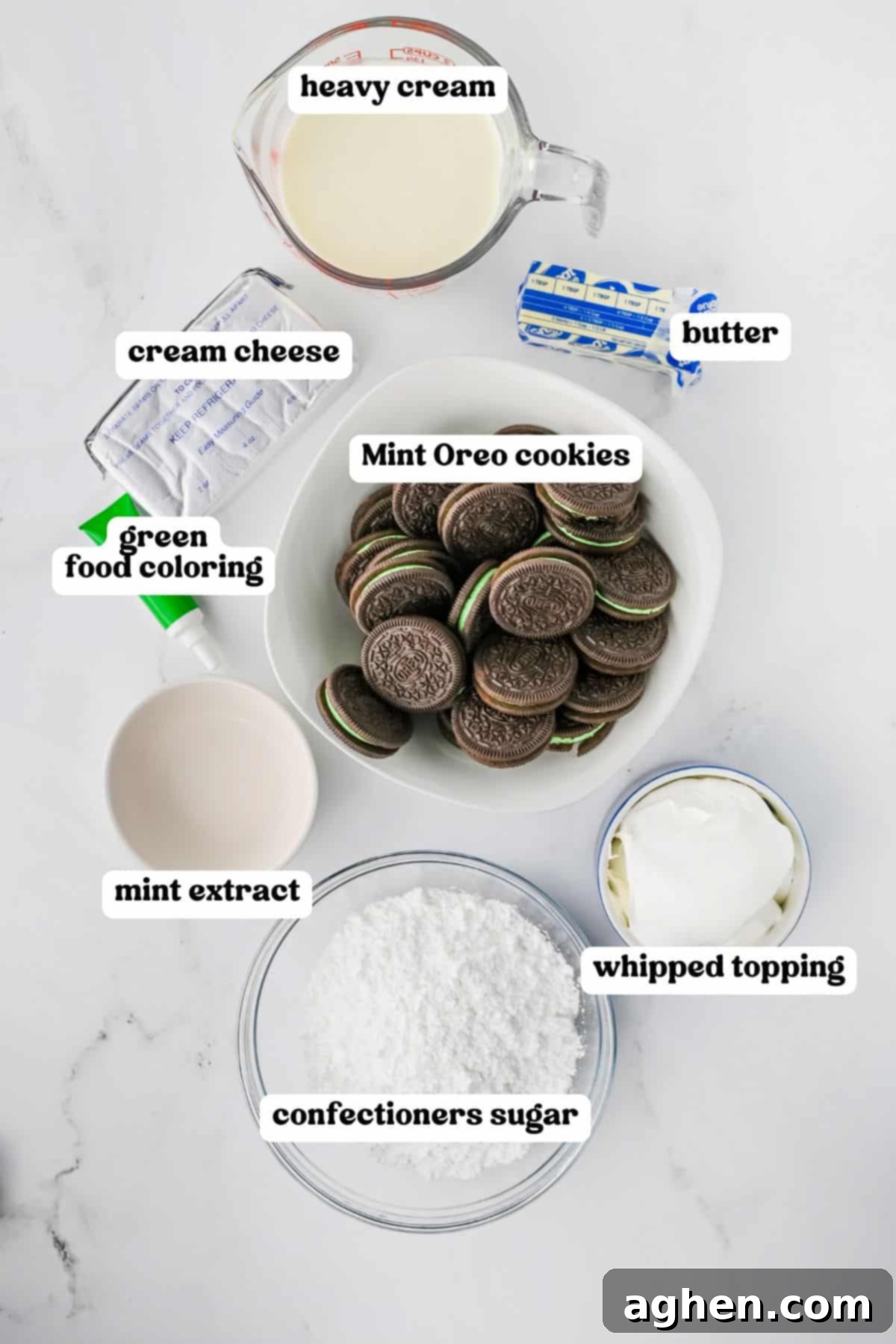A high-angle flat lay shot showcasing all the essential ingredients for No-Bake Grasshopper Mint Pie, neatly arranged on a white surface with clear labels. Ingredients include a carton of heavy cream, a block of cream cheese, a stick of butter, a bottle of green food coloring, a stack of Mint Oreo cookies, a bottle of mint extract, a tub of whipped topping, and a bowl of confectioners' sugar.