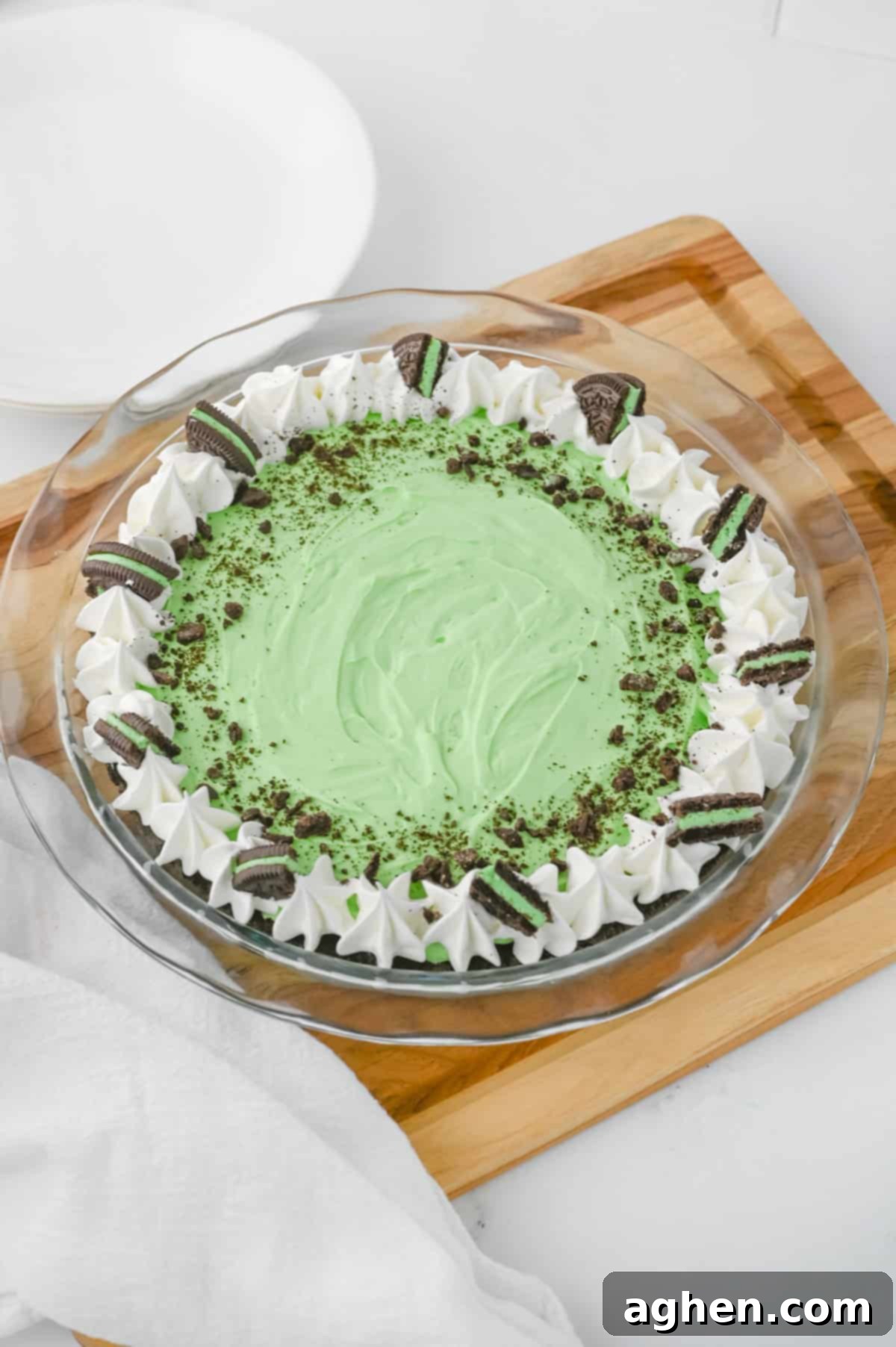The whole No-Bake Grasshopper Mint Pie presented beautifully in a clear glass pie dish. It features a vibrant green filling, topped with decorative swirls of whipped cream, a scattering of crushed chocolate cookies, and several Mint Oreo halves, creating an appealing and festive dessert.