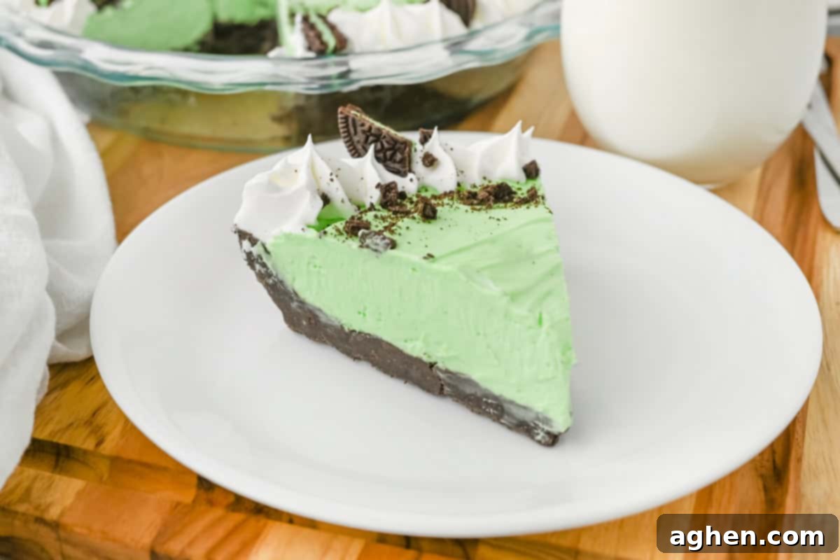 A close-up of a delectable slice of frozen mint Grasshopper pie, beautifully plated on a white dish. The vibrant green filling contrasts perfectly with the dark chocolate crust, and it's adorned with elegant swirls of whipped cream, a sprinkle of crushed cookies, and a whole Mint Oreo piece on top. In the background, the full pie in its glass dish and a refreshing glass of milk are visible.