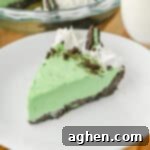A close-up of a slice of no bake grasshopper pie with green filling, chocolate crust, whipped cream swirls, crushed chocolate cookies, and a Mint Oreo half, served on a white plate.