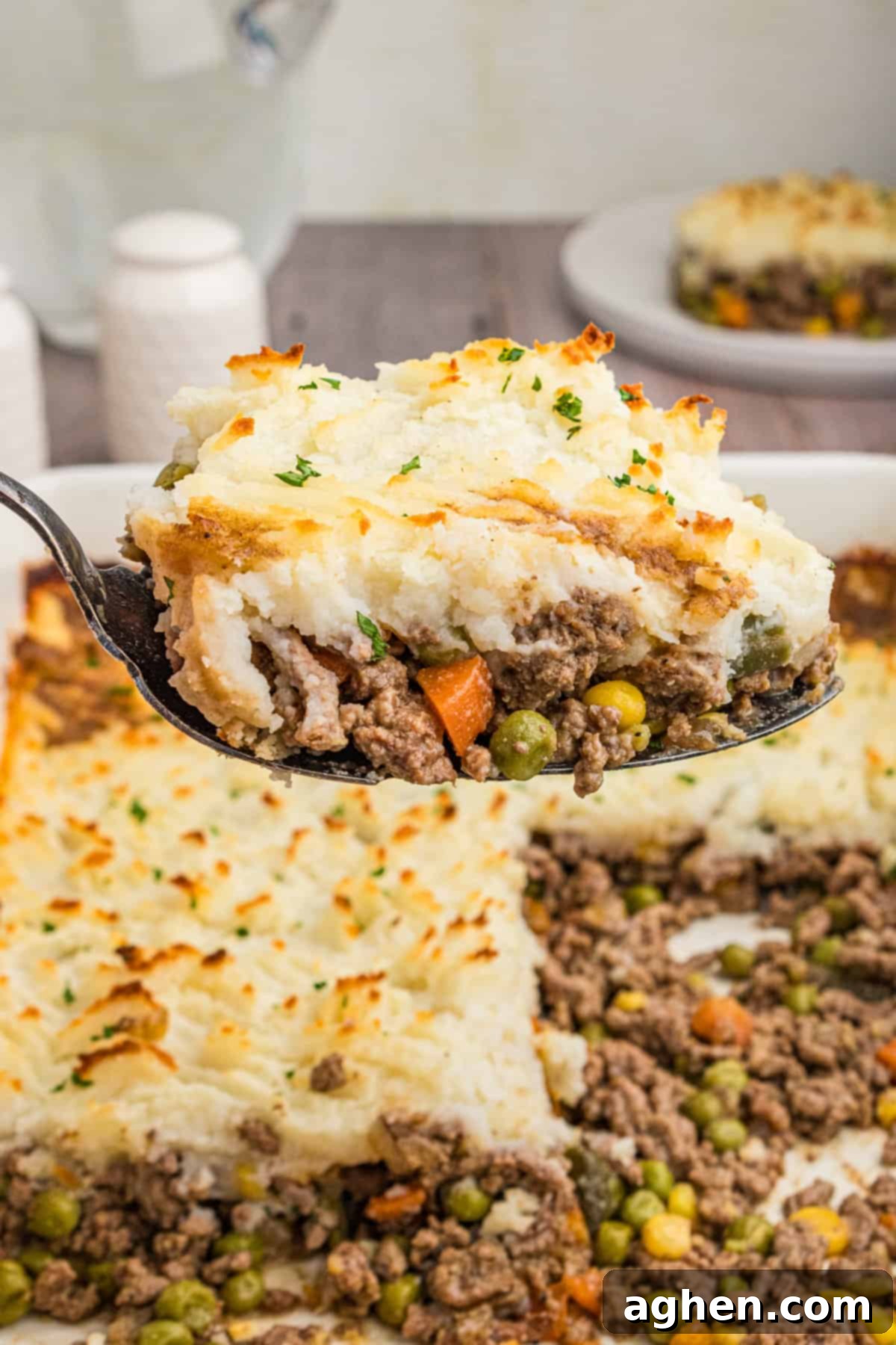 No-Fuss Cottage Pie 3 A close-up of a spoonful of cottage pie, showing golden mashed potatoes on top of a beef and vegetable mixture, garnished with chopped parsley. The casserole dish with the remaining pie is visible in the background.