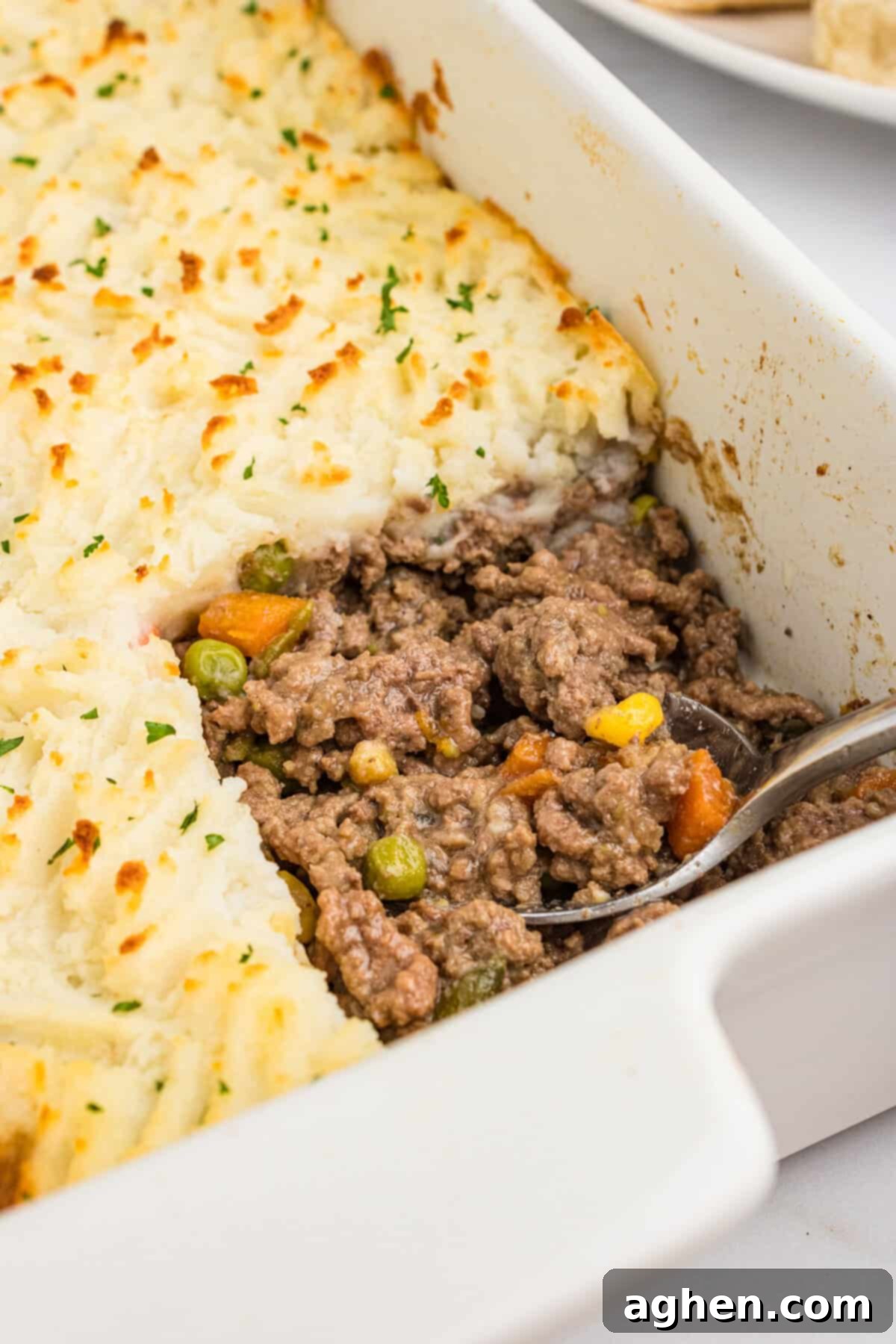 No-Fuss Cottage Pie 8 A close-up of a white casserole dish with a portion of cottage pie scooped out, revealing the seasoned ground beef, peas, carrots, corn, and green beans beneath the golden mashed potato topping.