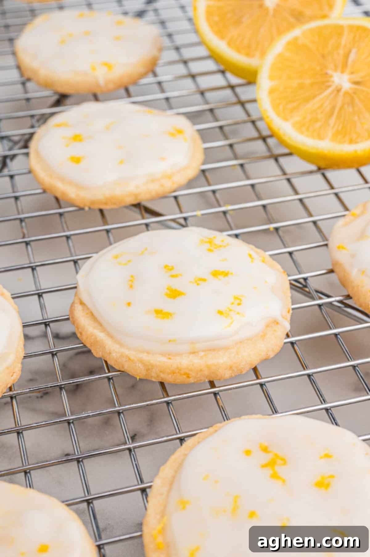 Several round lemon shortbread cookies with a smooth white glaze and sprinkled zest rest on a wire cooling rack, with sliced lemons in the background.