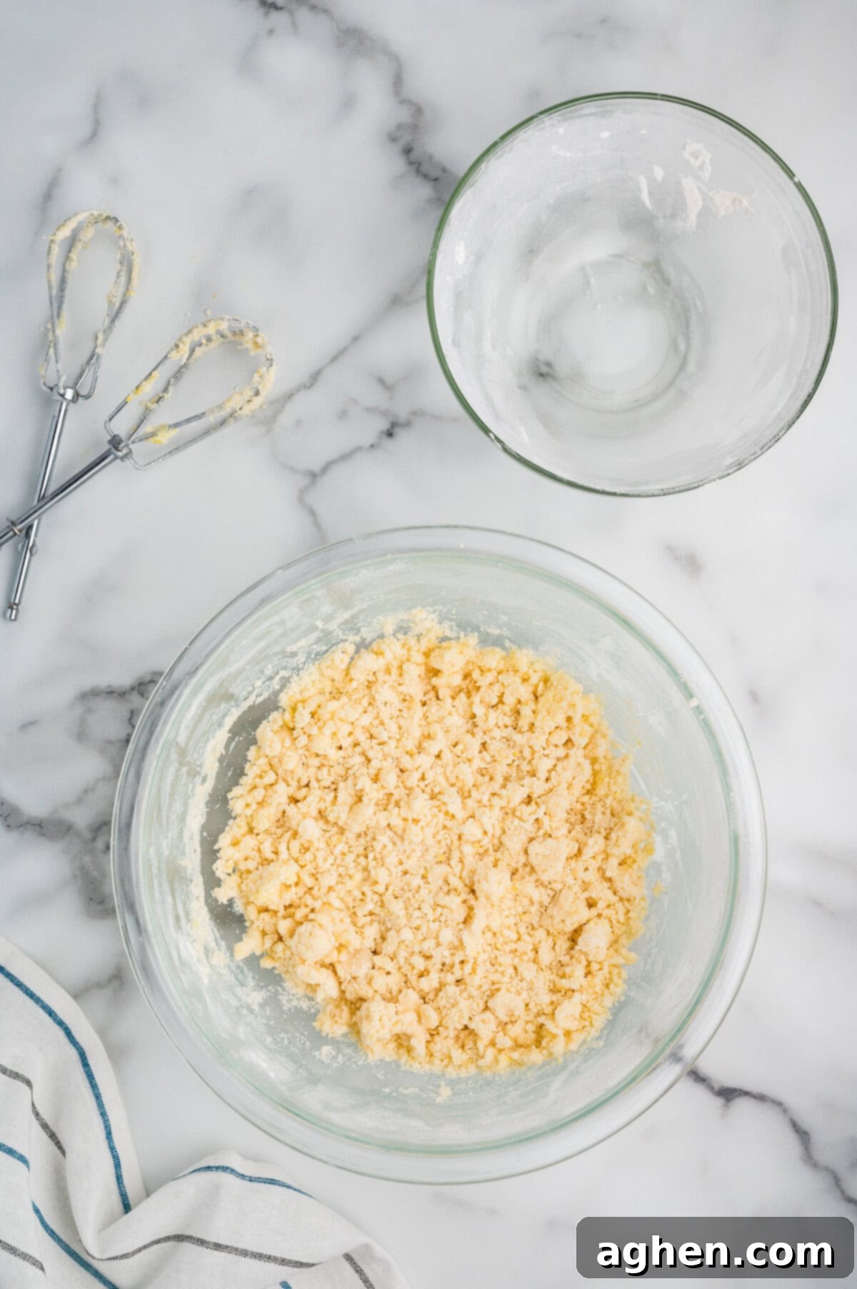 A glass bowl containing a crumbly shortbread dough mixture sits next to an empty flour bowl and butter-coated mixer beaters on a marble countertop.