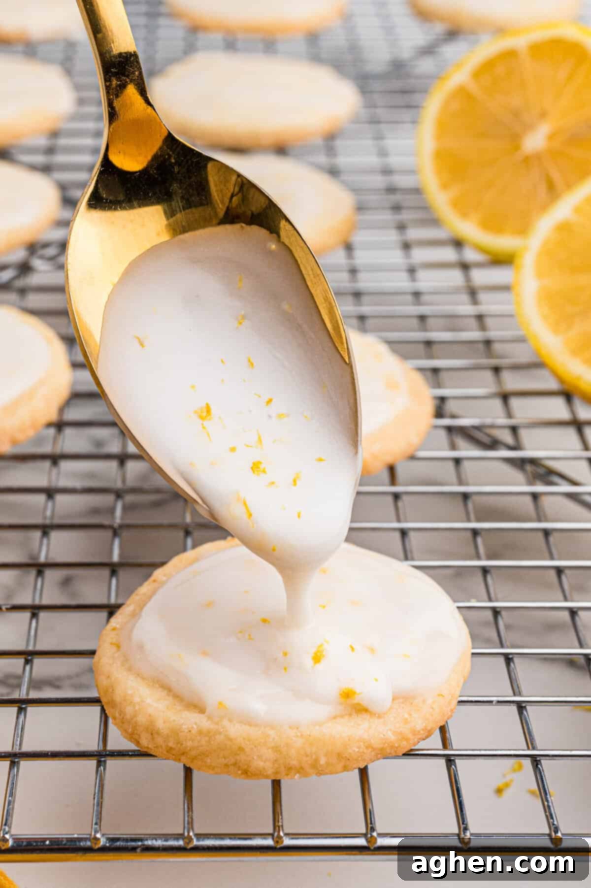 A close-up of a gold spoon drizzling thick white lemon glaze onto a round shortbread cookie, with zest speckled throughout.