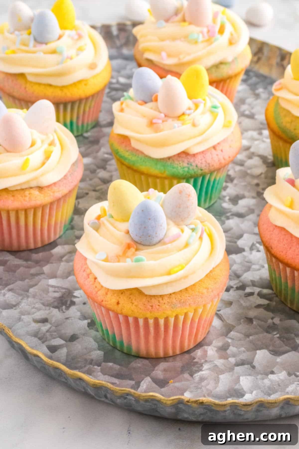Several pastel swirl Easter cupcakes sit on a round metal tray, each topped with piped buttercream frosting, pastel sprinkles, and mini chocolate eggs. 