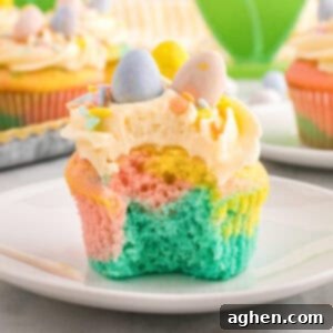 A close-up of a pastel swirl Easter cupcake with a bite taken out, revealing soft layers of blue, pink, and yellow cake. The cupcake is topped with piped buttercream frosting, pastel sprinkles, and three mini chocolate eggs in blue, yellow, and pink.