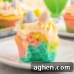 A close-up of a pastel swirl Easter cupcake with a bite taken out, revealing soft layers of blue, pink, and yellow cake. The cupcake is topped with piped buttercream frosting, pastel sprinkles, and three mini chocolate eggs in blue, yellow, and pink.