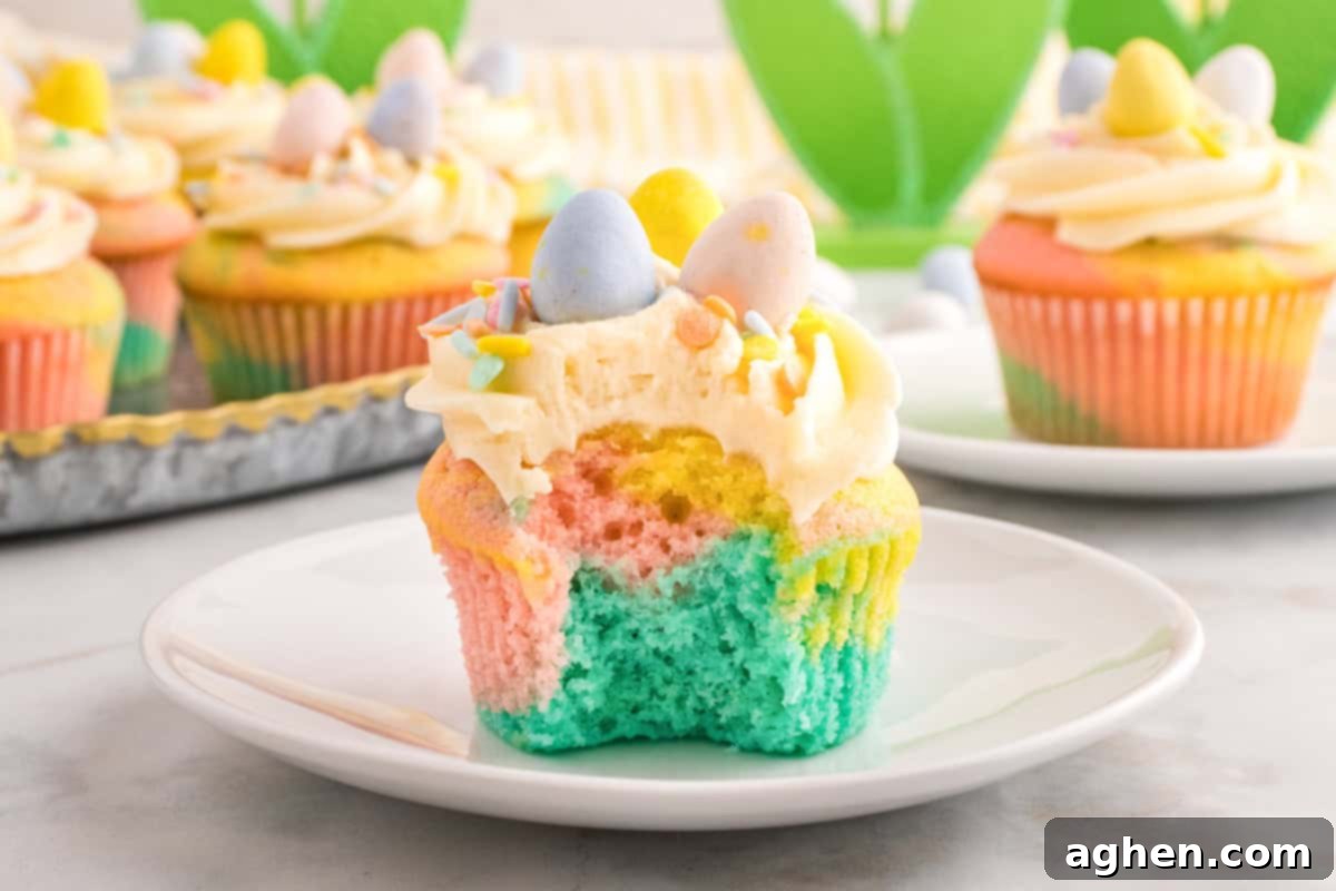 A close-up of a pastel swirl Easter cupcake with a bite taken out, revealing soft layers of blue, pink, and yellow cake. The cupcake is topped with piped buttercream frosting, pastel sprinkles, and three mini chocolate eggs in blue, yellow, and pink.