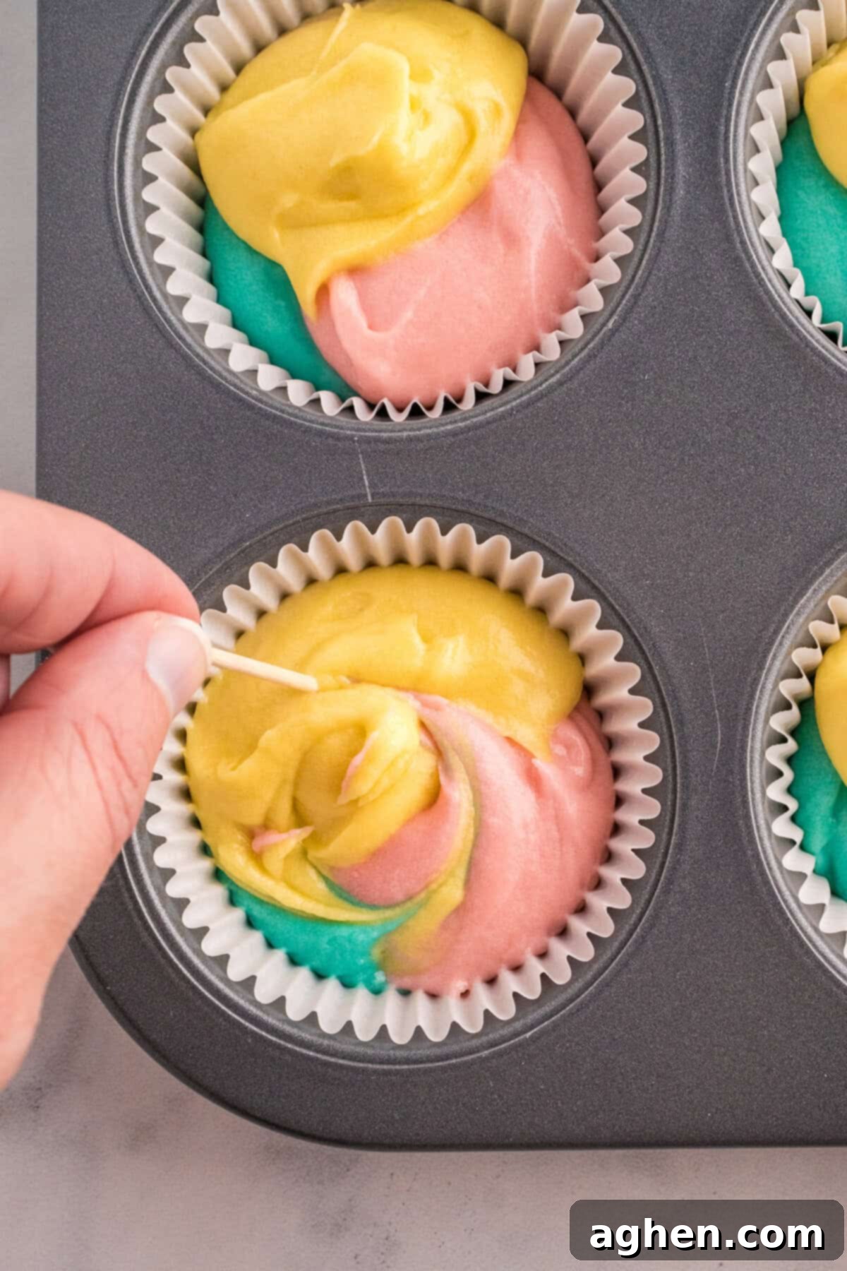 A hand using a toothpick to swirl yellow, pink, and blue cupcake batter inside a paper liner placed in a muffin pan. The colors blend slightly, creating a marbled effect before baking.