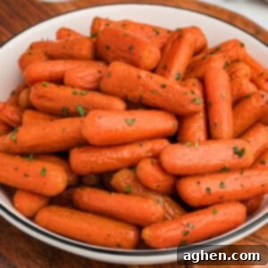 A close-up of a white bowl filled with glossy, brown sugar honey glazed baby carrots, garnished with finely chopped green herbs. The carrots have a slightly caramelized exterior and are arranged in a heaping pile.