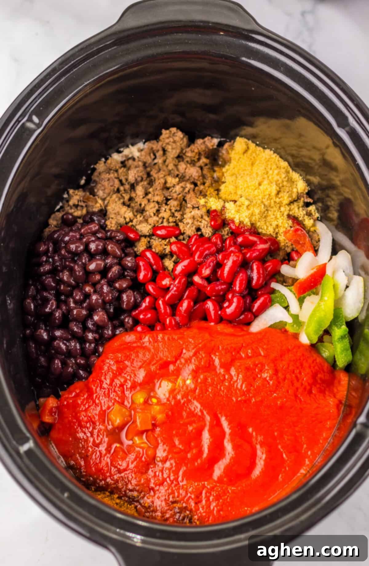 Top-down view of dr pepper chili ingredients in a slow cooker, showing separate piles of black beans, kidney beans, ground beef, tomato sauce, rotel, frozen peppers and onions, light brown sugar, and chili seasoning before mixing or cooking.
