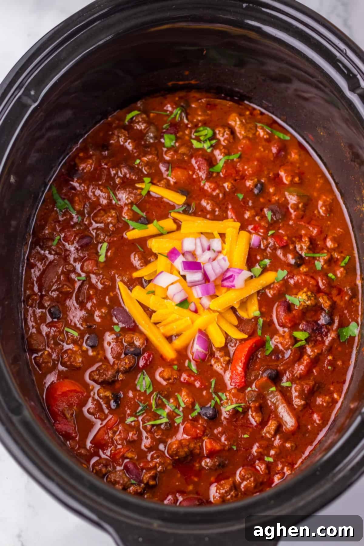 Thick and chunky Dr. Pepper Chili in a slow cooker with visible ground beef, black beans, kidney beans, peppers, and tomatoes. It’s garnished with shredded cheddar cheese, chopped red onions, and fresh herbs.