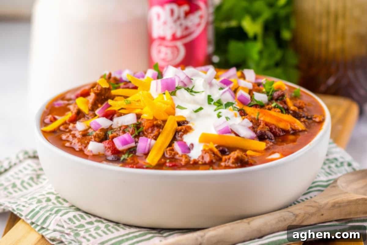 A side view of a white bowl filled with slow cooker dr pepper chili topped with shredded cheddar cheese, chopped red onions, sour cream, and fresh parsley. A can of Dr Pepper and fresh parsley are visible in the blurred background.

