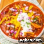 Bowl of Crockpot Dr Pepper Chili with ground beef, beans, and tomatoes in a red sauce, topped with shredded cheese, onions, and a dollop of sour cream.