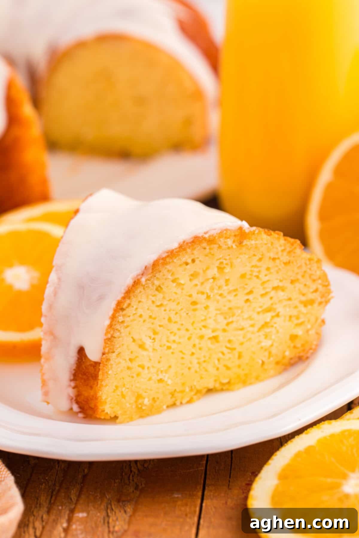 A slice of glazed orange juice cake with a smooth white icing glaze sits on a white plate. The background shows halved oranges, a glass of orange juice, and the rest of the orange bundt cake.