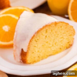 A slice of orange juice cake with a smooth white icing glaze sits on a white plate. The background shows fresh oranges.
