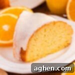 A slice of orange juice cake with a smooth white icing glaze sits on a white plate. The background shows fresh oranges.