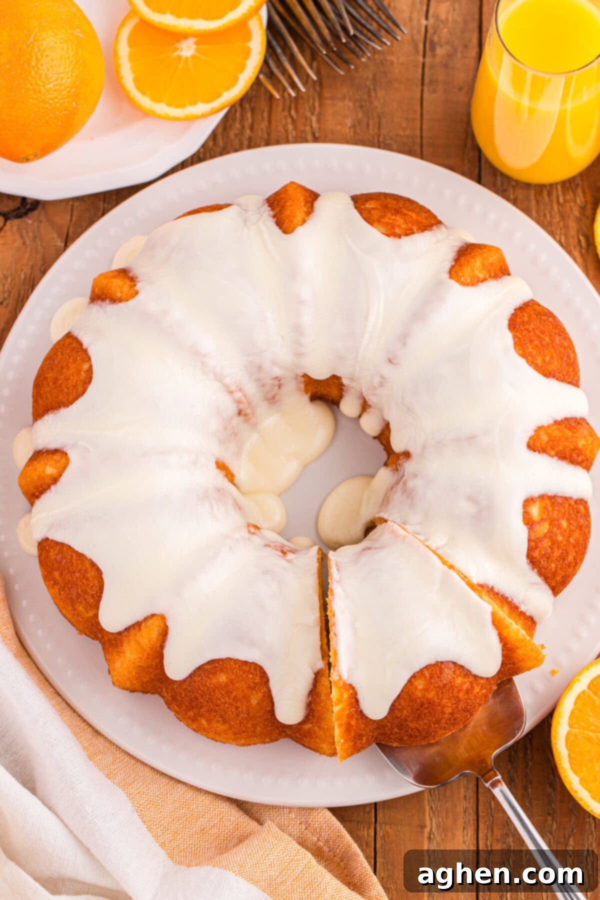An overhead view of a full orange juice bundt cake topped with white icing. A slice is being removed with a cake server, and orange slices, a glass of juice, and utensils are visible around the plate.