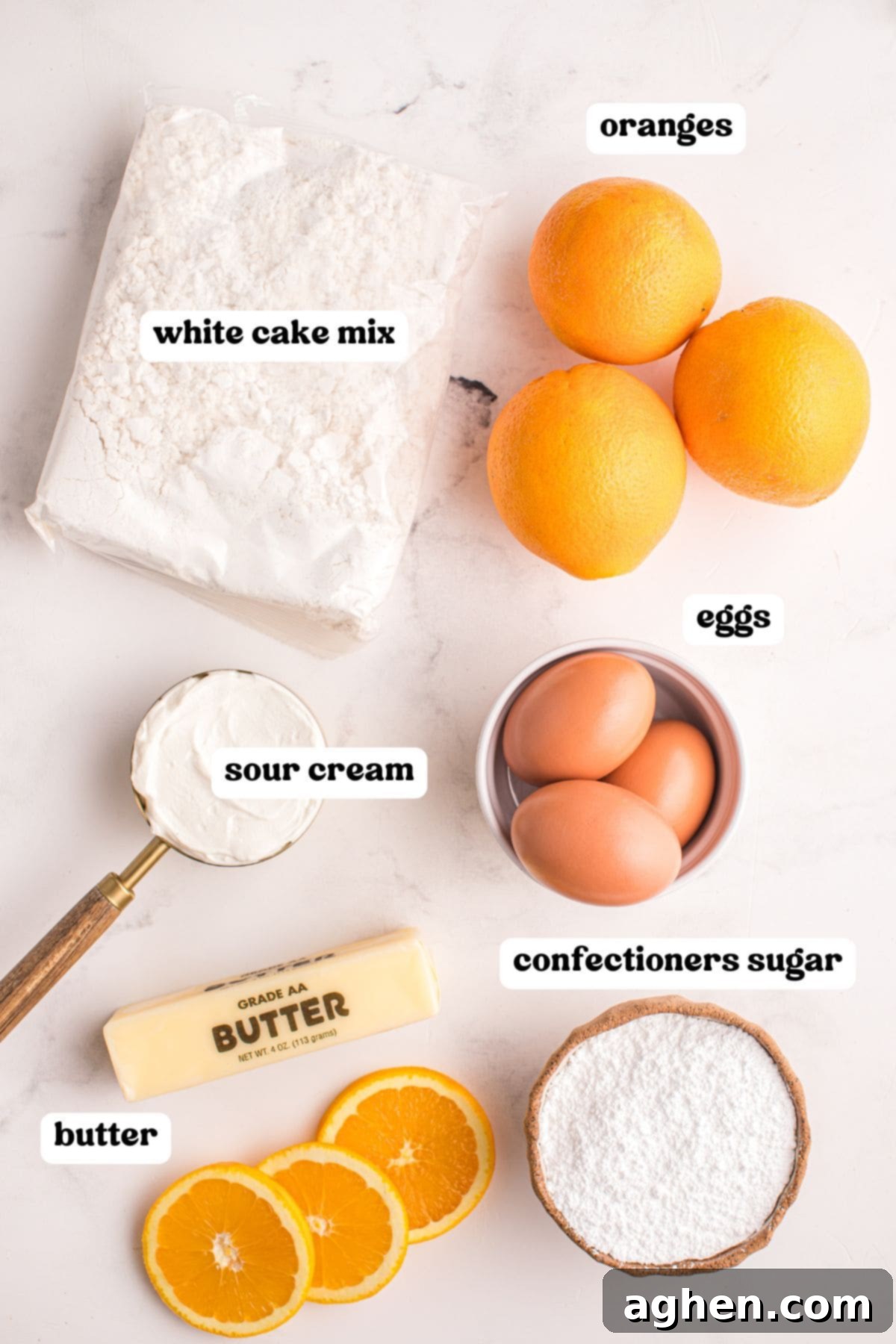 Flat lay of labeled ingredients for orange juice cake on a white surface, including a bag of white cake mix, three oranges, three eggs, confectioners sugar, a stick of butter, and sour cream.