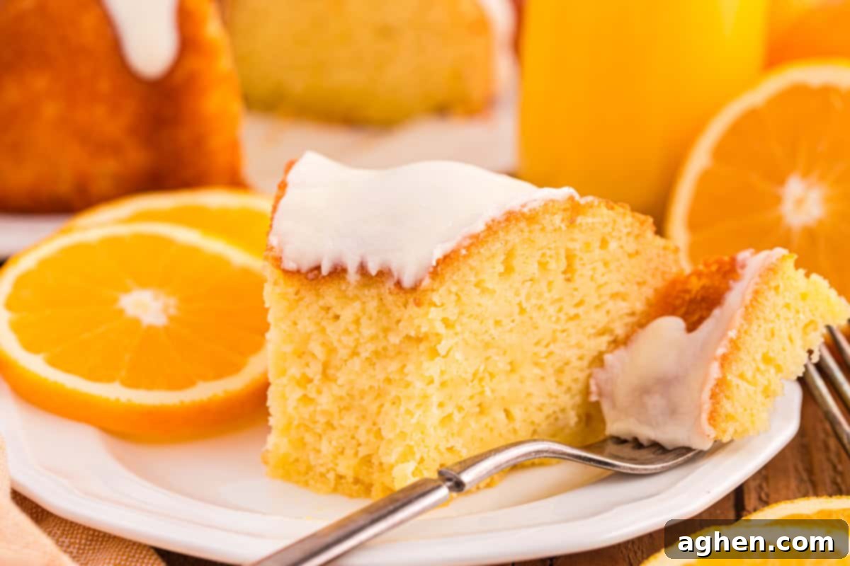 A close-up of a slice of moist orange juice cake topped with a layer of smooth white icing, served on a white plate with fresh orange slices. A fork holds a small bite of the cake.