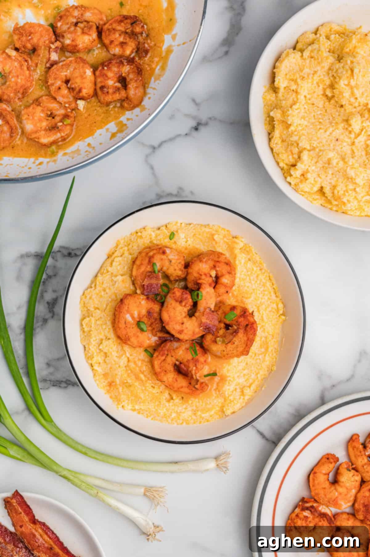 Overhead view of a bowl of cheesy shrimp and grits with bacon and green onions. Surrounding it are a skillet with saucy shrimp, a bowl of cheesy grits, a plate of cooked shrimp, fresh green onions, and a plate of bacon, all arranged on a marble surface.