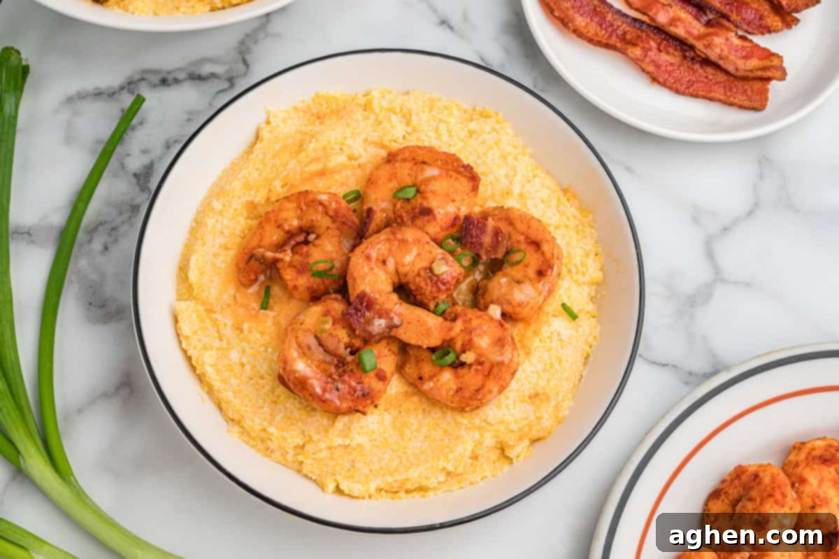 Bowl of cheesy grits topped with shrimp, bacon pieces, and scallions. The bowl is surrounded by fresh green onions and a plate of bacon.