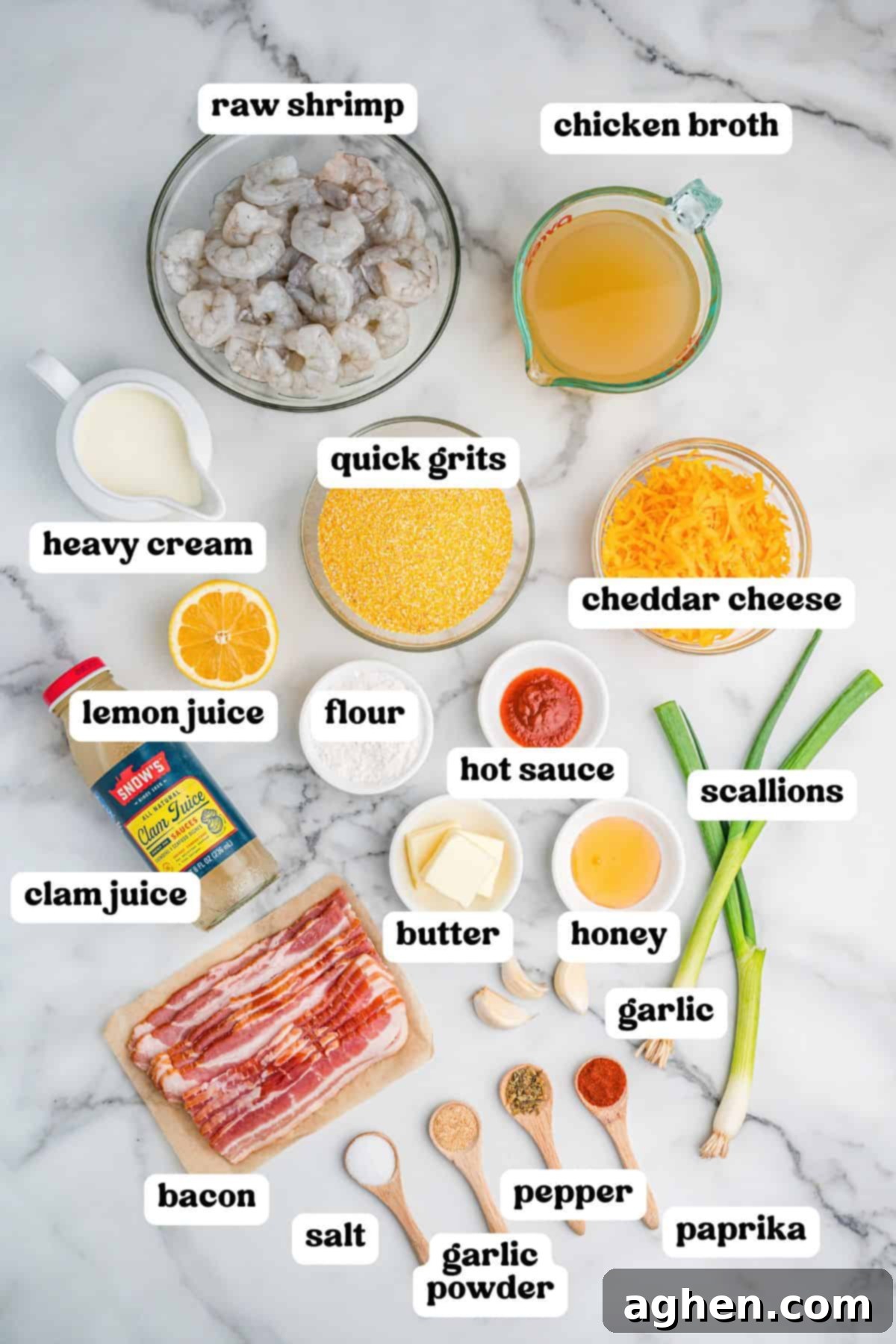 Overhead view of various labeled ingredients for cheesy shrimp and grits arranged on a marble surface. Items include raw shrimp, chicken broth, quick grits, cheddar cheese, heavy cream, lemon juice, flour, hot sauce, butter, honey, garlic, scallions, bacon, clam juice, and seasonings in wooden spoons: salt, garlic powder, pepper, and paprika.