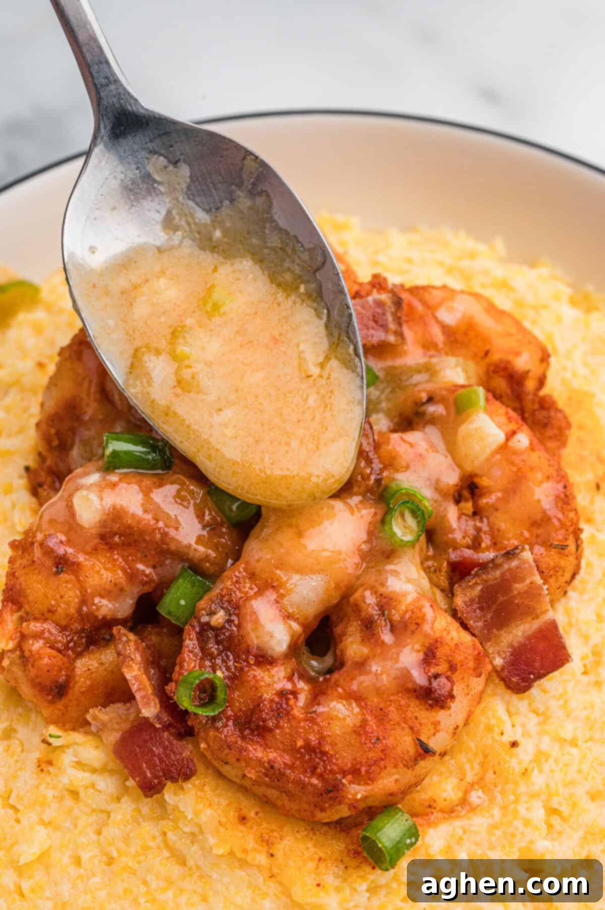 A spoon drizzles buttery sauce over a serving of shrimp on a bed of cheesy grits, garnished with bacon and green onions.