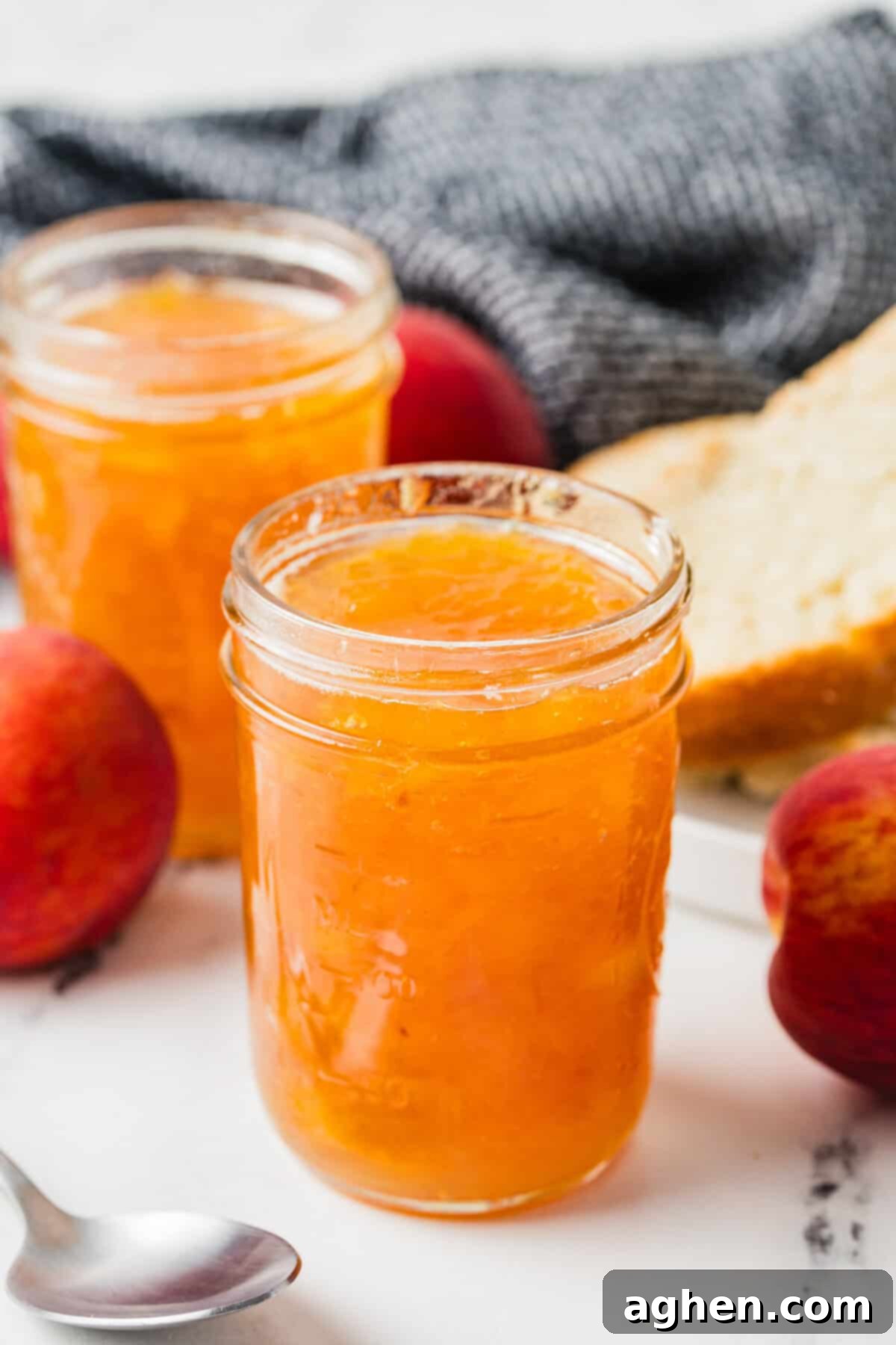 Two jars of homemade peach jam with whole peaches, a spoon, and a plate with sliced bread in the background. Freshly made small-batch peach jam, perfect for toast and breakfast.